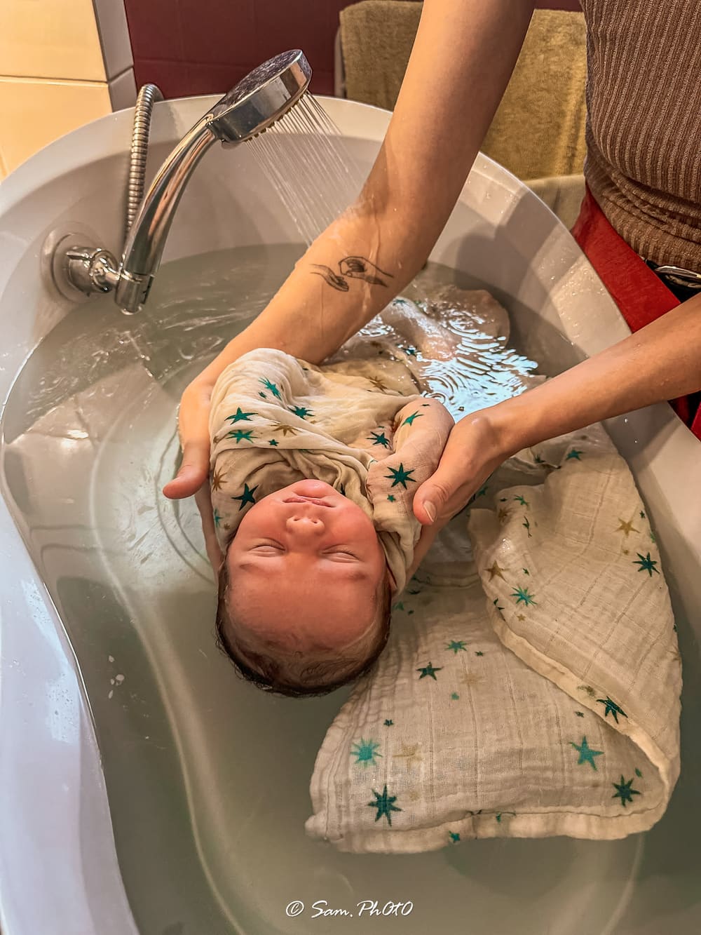 Aurore Lucas réalisant un bain enveloppé à un nouveau-né emmailloté dans un lange étoilé, sous un filet d'eau tiède